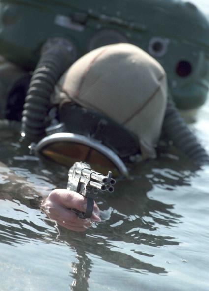 Водолаз выныривает из воды, в руках у него подводный пистолет с четырьмя стволами.