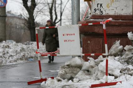 На углу дома стоит табличка с надписью- ОСТОРОЖНО СОСУЛЬКИ мимо проходит женщина. 3 марта 2011 года.