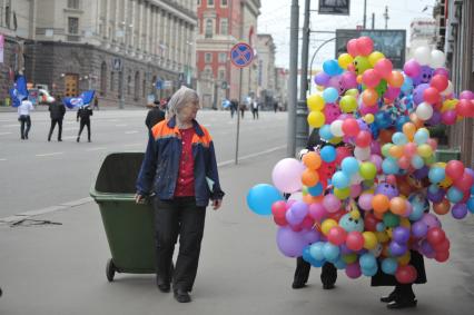 На 1 мая на Тверской женщина- гастарбайтер идет по улице с мусорной тележкой мимо места продажи воздушных шаров, Москва, 1 мая 2011 года