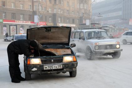 Поломанный автомобиль стоит посреди улицы. Мужчина пытается его починить.