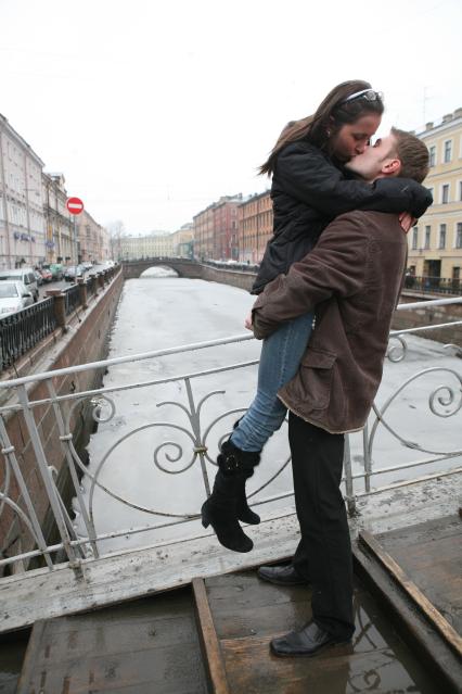 Молодая пара стоит на мосту и целуются. Мужчина поднял женщину на руки.