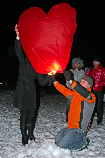 Мужчина и женщина держат в руках китайский летающий фонарь в виде сердца. Мужчина поджигает свечку.