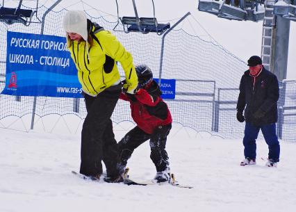 Молодая мама выкатывает за руки своего ребенка в начало спуска во время обучения катанию на горных лыжах.