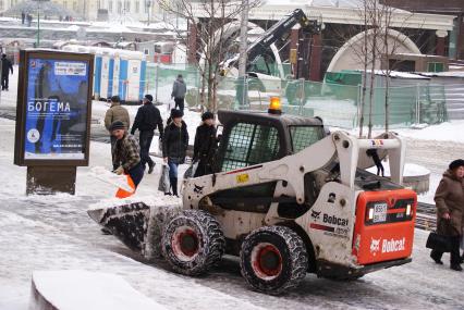 Снегоуборочная техника. Рабочий закидывает лопатой снег в ковш мини-экскаватора.