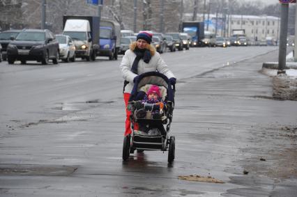 Молодая женщина с ребенком в детской коляской идет вдоль дороги.