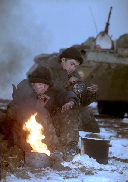 На переднем плане двое солдат в грязной военной форме сидят у огня разведенного в военной каске.
На заднем плане - Боевая Машина Пехоты.