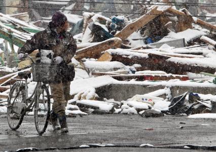 Сильнейшее землетрясение и цунами в Японии. Последствия разрушений. Мужчина с велосипедом идет мимо разрушенного строения.