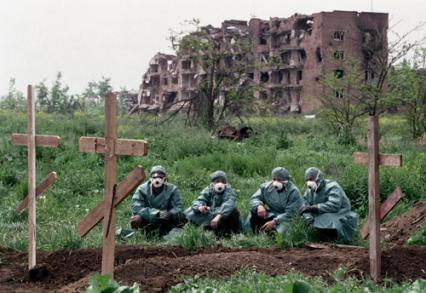 Похоронщики в респираторах сидят рядом со свежими могилами недалеко от разрушенного дома в Чечне.