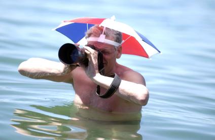 Фотограф снимает стоя в воде с зонтиком на голове.