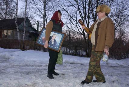 Мужчина с гитарой и бутылкой водки и женщина с портретом Владимира Путина стоят на улице в деревне зимой.