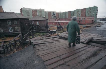 Пожилая женщина идет по дороге мимо старых и новых высоких домов.