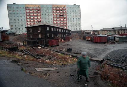 Пожилая женщина идет по городу. Старый двухэтажный дом стоит на фоне высокой новостройке.