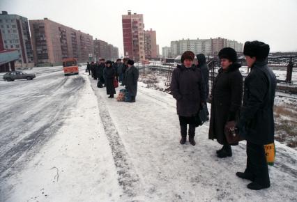 Зима. Люди стоят на остановке в районе с высокими домами.