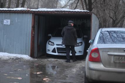 Мужчина стоит у открытого металлического гаража во дворе дома.