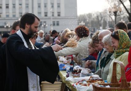 На улице священник освящает куличи, бутылки вина и крашенные яйца обрызгивая кропилом стол с угощениями и прихожан.