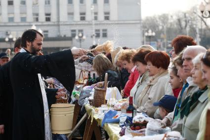 На улице священник освящает куличи, бутылки вина и крашенные яйца обрызгивая кропилом стол с угощениями и прихожан.