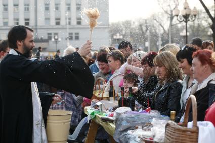 На улице священник освящает куличи, бутылки вина и крашенные яйца обрызгивая кропилом стол с угощениями и прихожан.