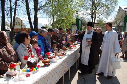 На улице священник освящает куличи, бутылки вина и крашенные яйца обрызгивая кропилом стол с угощениями и прихожан.