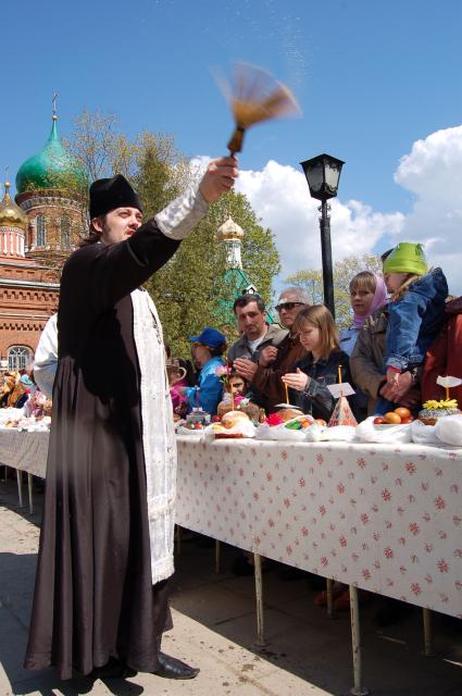 На улице священник освящает куличи, бутылки вина и крашенные яйца обрызгивая кропилом стол с угощениями и прихожан.