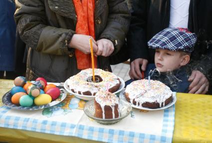 На улице, около стола на котором стоят пасхальные куличи и крашенные яйца стоит ребенок.