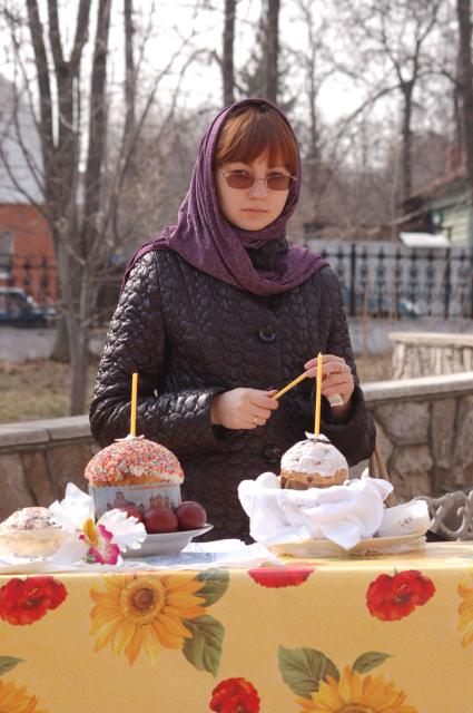 Молодая девушка стоит у стола с пасхальными куличами и крашенными яйцами, держит в руках свечку.