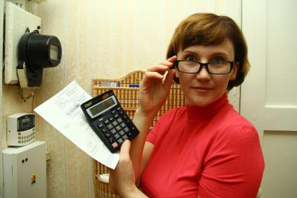 Женщина в очках с калькулятором и квитанцией на оплату коммунальных услуг стоит около счетчика электроэнергии.