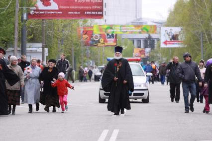 Священник с гвоздикой и георгиевской лентой идет по улице города. Сзади едет автомобиль полиции.