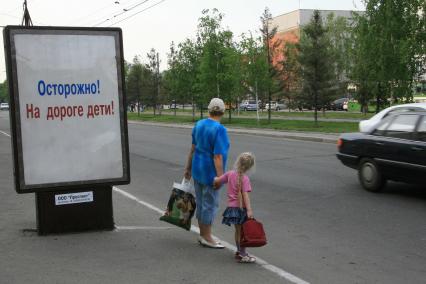 Бабушка с внучкой собираются нарушить правила дорожного движения перейдя автомобильную дорогу в неположенном месте. Рядом стоит рекламный щит с социальной рекламой `Осторожно! На дороге дети!`.