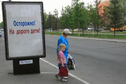 Бабушка с внучкой собираются нарушить правила дорожного движения перейдя автомобильную дорогу в неположенном месте. Рядом стоит рекламный щит с социальной рекламой `Осторожно! На дороге дети!`.