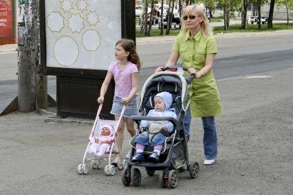 По улице города идет мама с двумя детьми. Один ребенок в коляске, второй везет коляску с куклой.