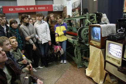 Краеведческий музей. Посетители смотрят телевизионные программы времен СССР по черно-белым телевизорам. Сзади плакат с надписью: `Иди, товарищ, к нам в колхоз!`.