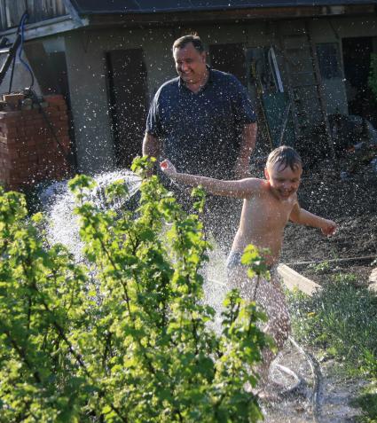 Ребенок на даче бежит от папы, который поливает его водой из шланга.