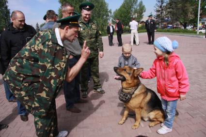 Мужчина в форме пограничника что-то рассказывает собаке с намордником, которую за ухи трепят двое смеющихся детей.
