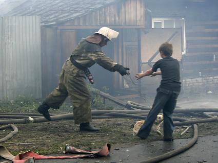 Пожарный за поводок передает собаку мальчику на фоне горящего дома.