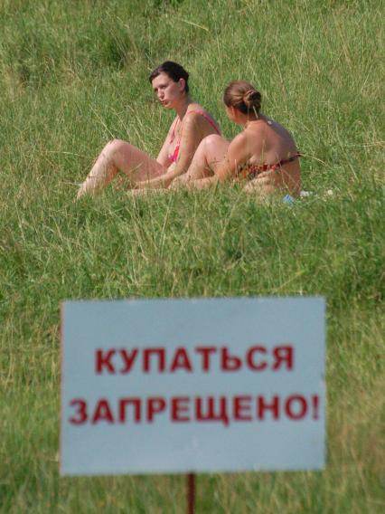 Девушки сидят в траве рядом с табличкой: `Купаться запрещено!`.