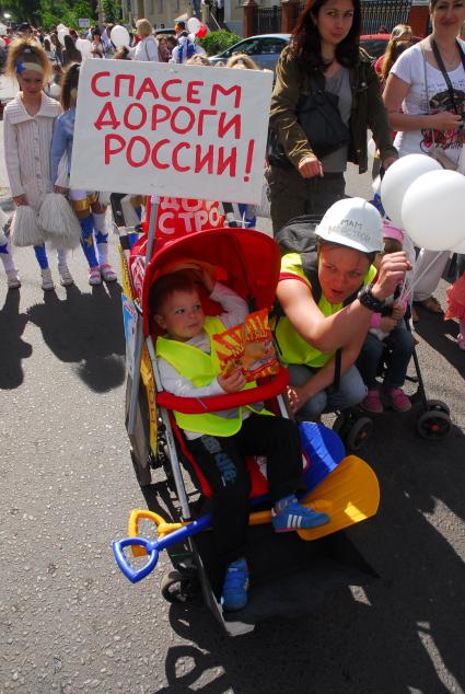 Мальчик одетый в одежду ремонтника в детской коляске. На коляске табличка с надписью: `Спасем дороги России!`