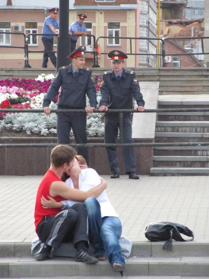 Молодая пара целуется на улице города, сзади стоят полицейские следящие за порядком.