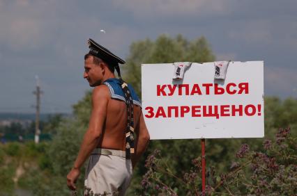 Молодой человек в бескозырке рядом с объявлением о запрете купания.