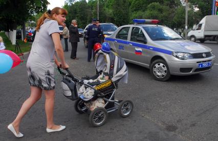 Мамаша везет детскую коляску стилизованную под полицейский автомобиль. Рдом стоит полицейский автомобиль с представителями полиции.