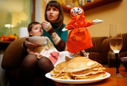 Девушка кормит ребенка на фоне тарелки с блинами и чучела масленицы.