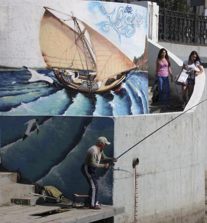 Международный фестиваль граффити в Казани посетили художники из Испании, Германии и Польши. На снимке: мужчина ловит рыбу на набережной, по лестнице спускаются две девушки, стена набережной оформлена рисунком корабля.