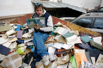 Старые книги лжат в мусорном контейнере возле университета РГУ им. Канта в Калининграде. На свалке сидит мужчина и читает одну из выброшенных книг.