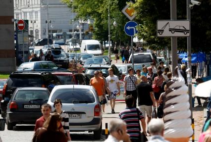 Хаотично расставленные припаркованные автомобили на улице города, мимио проходят люди. Висит знак предупреждающий о работе эвакуаторов.