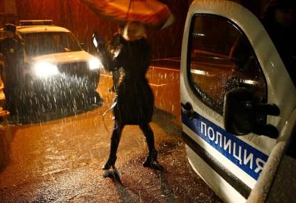 В дождливую погоду на месте происшествия в свете фар полицейского автомобиля идет работа. Девушка под зонтом с микрофоном в руках.