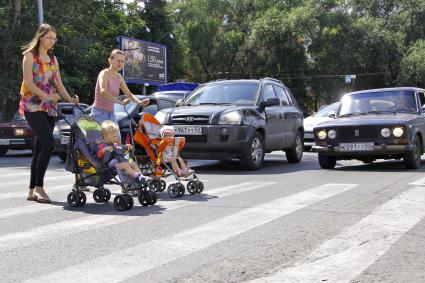 Девушки с колясками переходит автомобильную дорогу по пешеходному переходу.