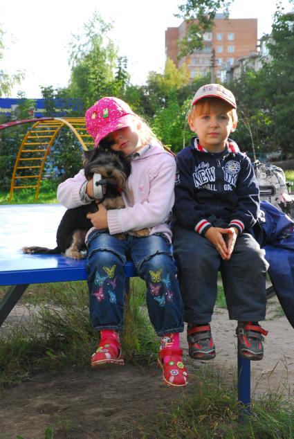 Мальчик и девочка сидят рядом во дворе. Девочка обняла собаку.