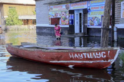 Летний паводок на реке Обь. Лодка с названием `Наутилус` пришвартована у магазина. Женщина в резиновых сапогах идет по воде.