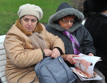 Две немолодые женщины сидят в парке на скамейке, одна из них рассматривает журнал с рекламой нижнего белья.