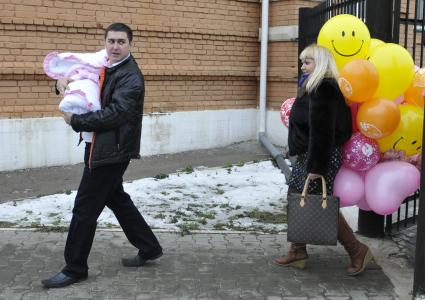 Высписка новорожденного с мамой из роддома. На снимке: Отец несет ребенка в конверте сзади идет женщина с воздушными шарами.
