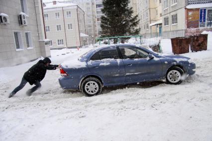 Мужчина толкает застрявший в снегу автомобиль.
